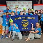 A group of students and adults wearing blue MESA shirts hold an Oregon state flag and pose, some wearing green hats, at a National Engineering Design Competition event. A Washington state flag is also visible.