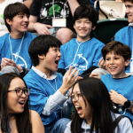 A group of smiling children in blue T-shirts sit closely together in stadium seats, laughing and talking enthusiastically at what appears to be a science or tech event like MESA Day 2026.