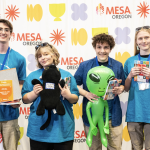 Four smiling students in blue shirts stand in front of a MESA Oregon backdrop, holding certificates, a black cat plush, an inflatable green alien, and boxes of prizes.