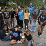 A group of smiling kids and two adults pose together outdoors on a sidewalk, with greenery in the background. Some are standing, while others are sitting or kneeling at the front.